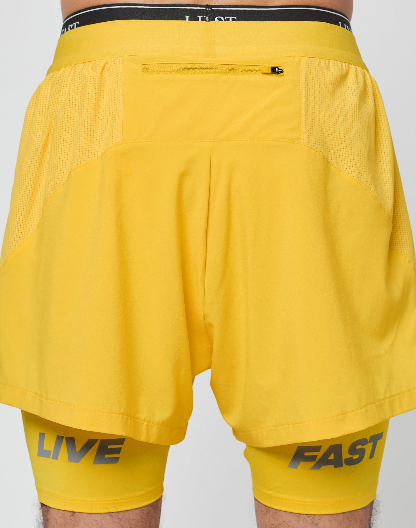 A person wears LIVE FAST DIE YOUNG Live Fast Performance Running Shorts in bright yellow, featuring lightweight fabric, a zippered back pocket, and integrated tights with LIVE and FAST printed in gray near each hem.