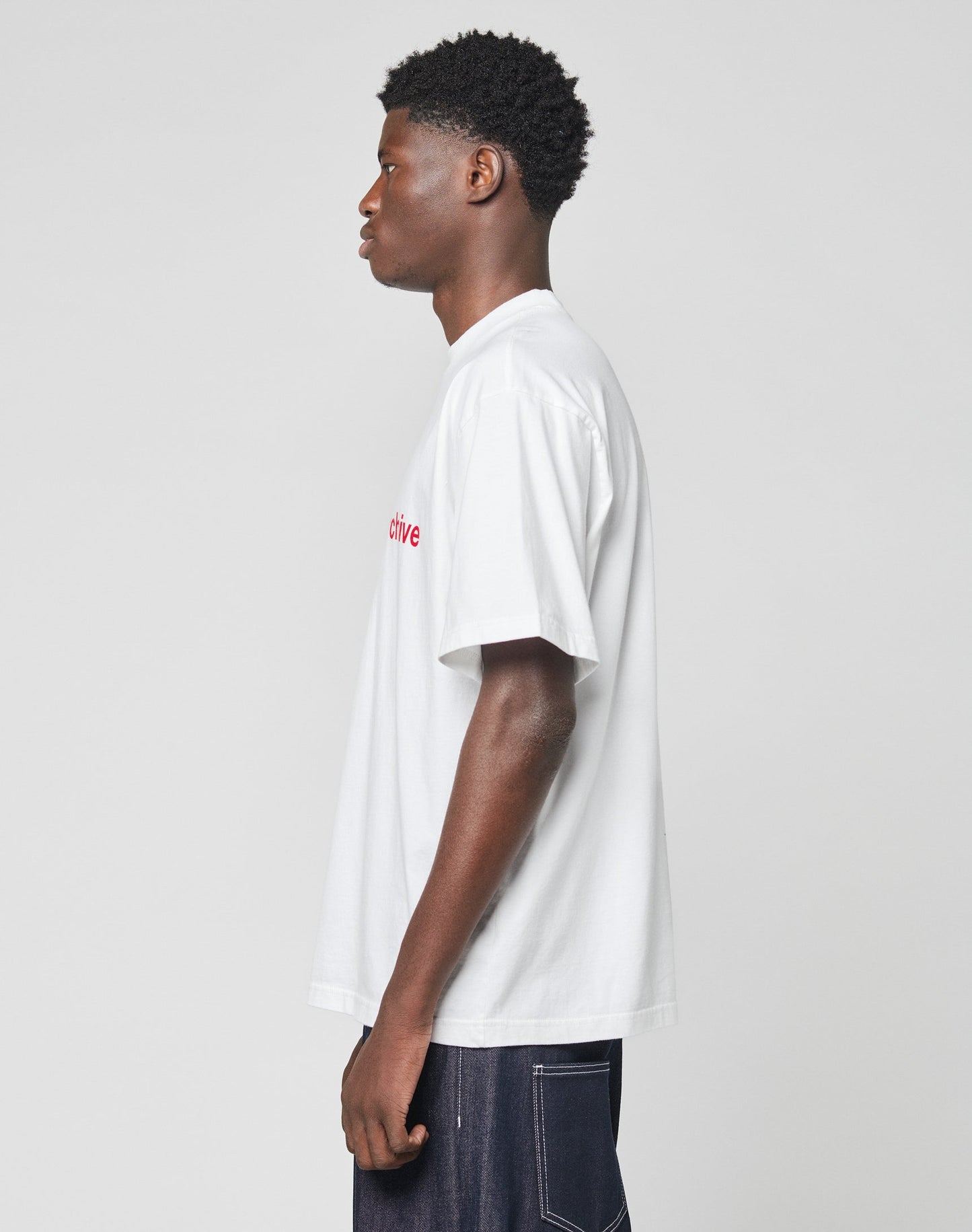 A man stands in profile against a plain background, wearing the Live Fast Archive Tee by LIVE FAST DIE YOUNG with red text and dark jeans.