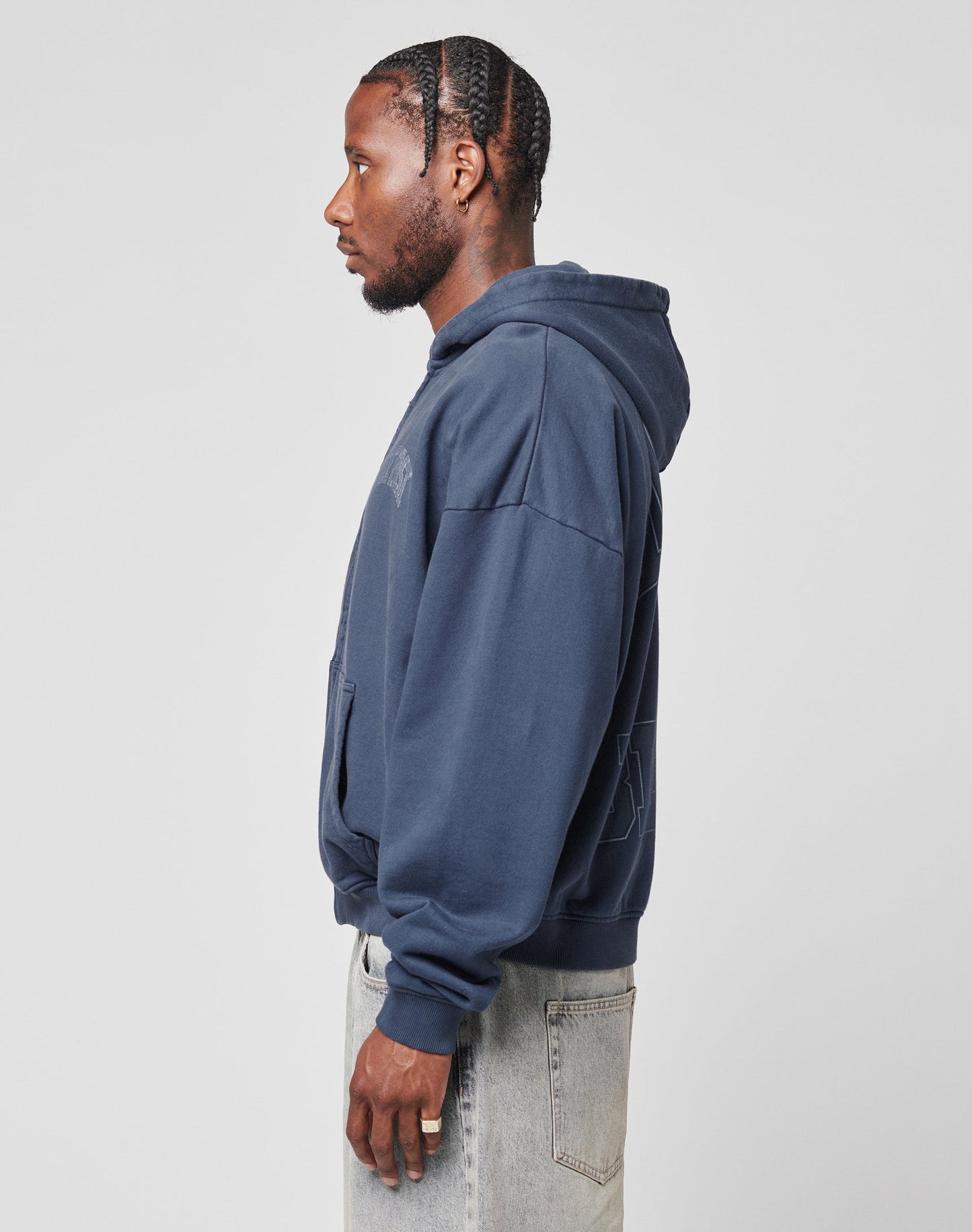 A man in profile wears the LIVE FAST DIE YOUNG Live Fast Berlin Hooded Zip in navy blue, styled in an oversize fit, paired with light-wash jeans. He has neat cornrows, a beard, and earrings, posing against a plain light gray background.