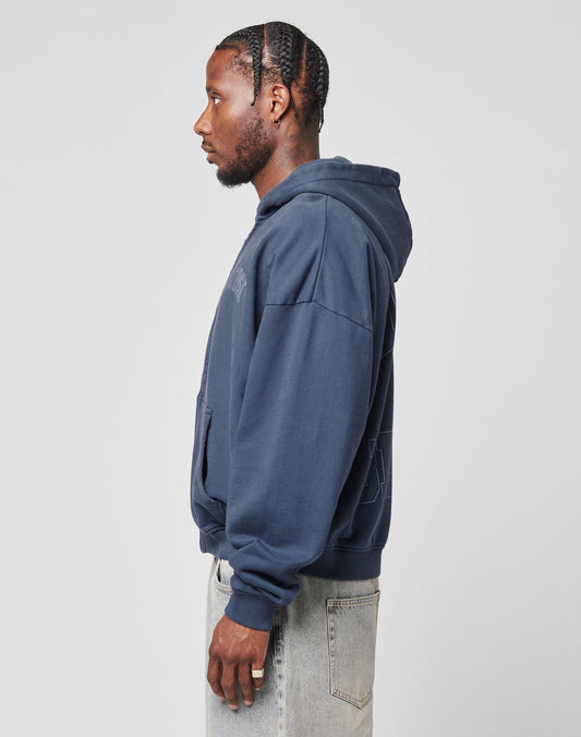 A man in profile wears the LIVE FAST DIE YOUNG Live Fast Berlin Hooded Zip in navy blue, styled in an oversize fit, paired with light-wash jeans. He has neat cornrows, a beard, and earrings, posing against a plain light gray background.
