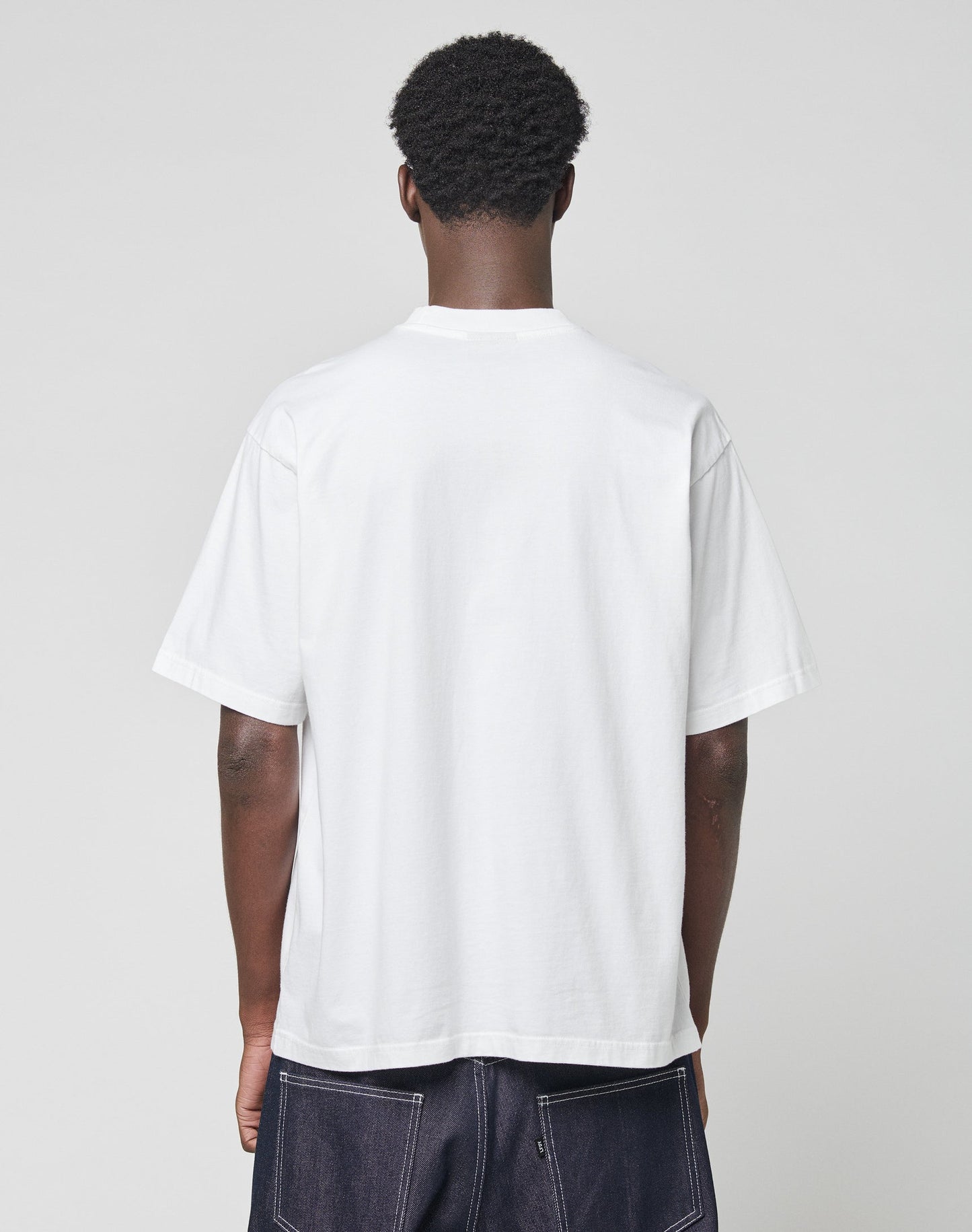 A person with short, curly hair stands with their back to the camera, wearing a plain white Live Fast Archive Tee by LIVE FAST DIE YOUNG in Oversize Fit and dark denim jeans against a light gray background.