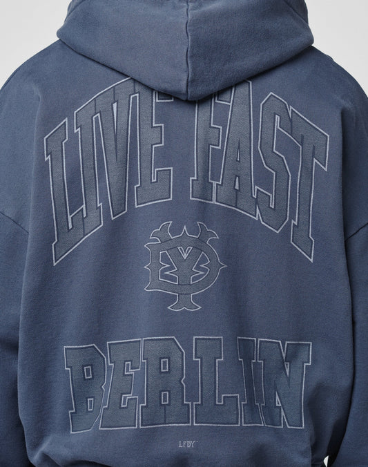 A person wearing the LIVE FAST DIE YOUNG Live Fast Berlin Hooded Zip in blue Baumwolle, oversize fit, with a vintage wash, bold LIVE FAST and BERLIN text plus graphic emblem and small LFDFY on the back.