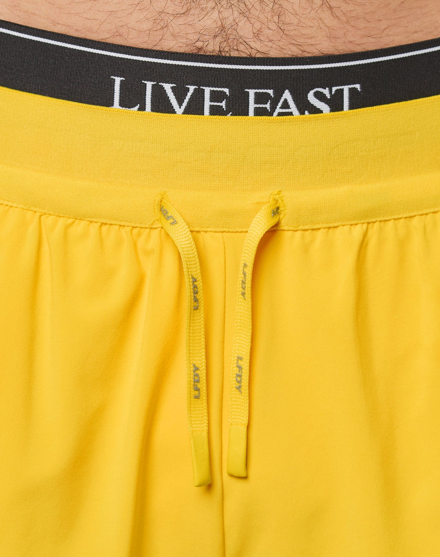 Close-up of LIVE FAST DIE YOUNG branding on the waistband of Live Fast Performance Running Shorts in bright yellow, featuring matching yellow drawstrings and partial view of a persons lower abdomen.
