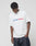 A man wearing the LIVE FAST DIE YOUNG Live Fast Archive Tee, an oversize white cotton T-shirt with Live Fast Archive in blue and red, stands against a plain background with his head down and one hand in his pocket.