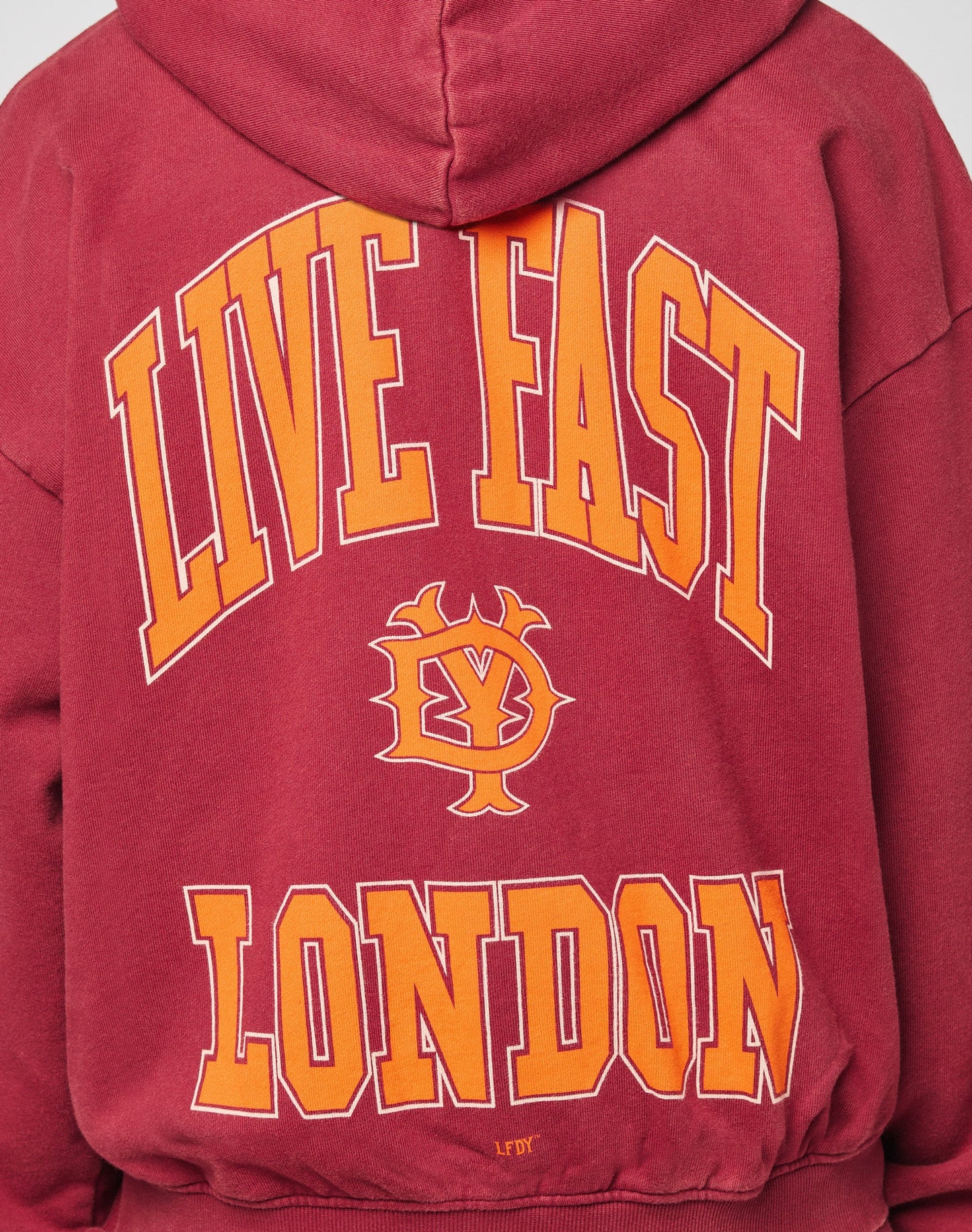 A person is seen from behind wearing an oversize fit maroon Live Fast London Hooded Zip by LIVE FAST DIE YOUNG, featuring yellow and orange LIVE FAST LONDON text and a small central graphic.| model