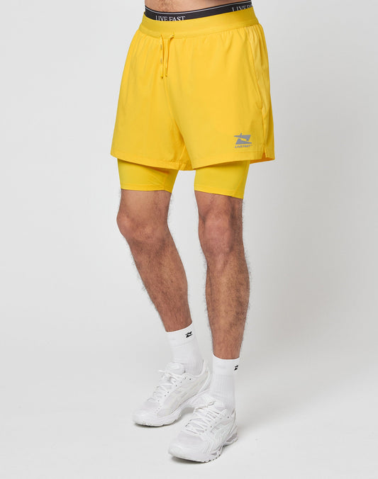 A person wears LIVE FAST DIE YOUNG Live Fast Performance Running Shorts with integrierte Tights, leichtes Obermaterial, and Live Fast branding on the waistband, paired with white socks and sneakers against a plain background.