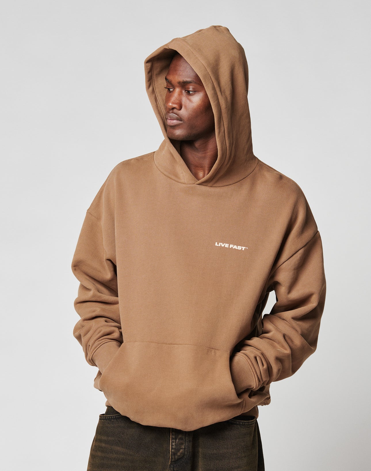 A person in an oversize fit brown Live Fast Plain Hooded sweatshirt by LIVE FAST DIE YOUNG, made from Baumwolle and Polyester with LIVE FAST. on the chest, stands hands-in-pocket looking to the side against a plain light background.