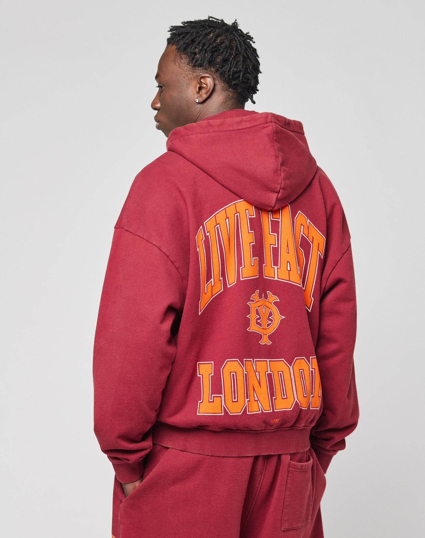 Close-up of the Live Fast London Hooded Zip by LIVE FAST DIE YOUNG in maroon oversize fit, featuring bold orange and yellow LIVE FAST lettering on the chest. Only part of the upper body is visible; crafted from soft Baumwolle.