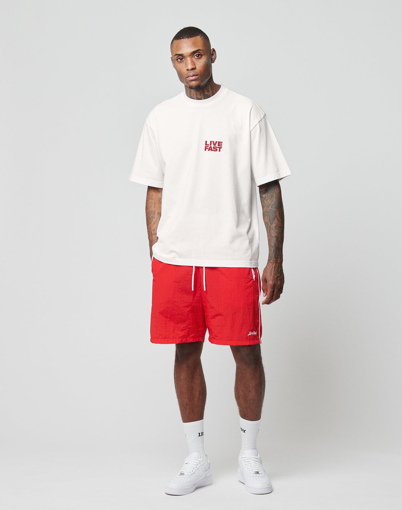 A man stands against a plain background wearing the LIVE FAST DIE YOUNG LFDY 4 Ever Tee, featuring an oversize fit, white cotton fabric, and front and back prints. He pairs it with red shorts, white socks, and sneakers, looking at the camera with hands in pockets.