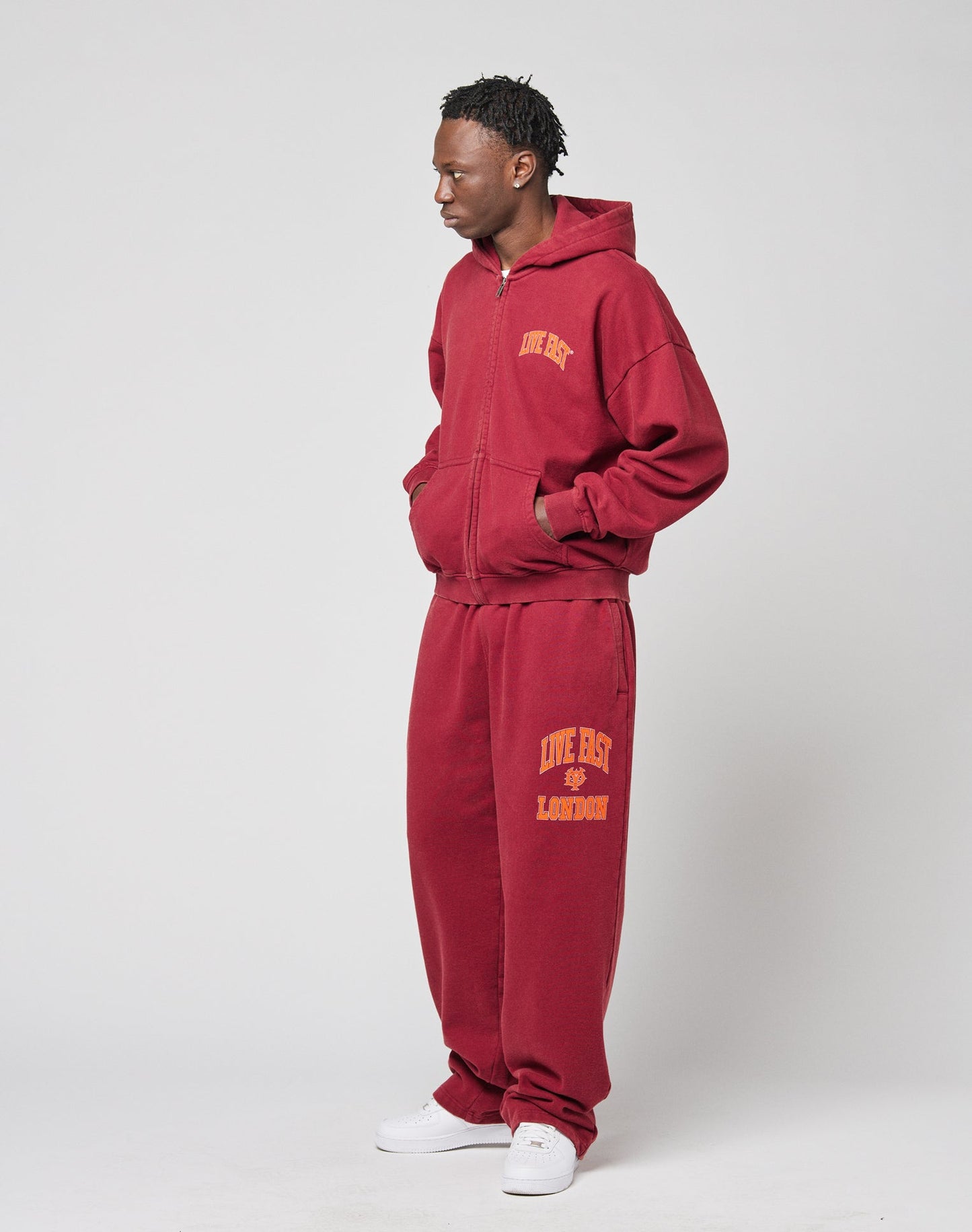 A man stands against a plain background wearing the LIVE FAST DIE YOUNG “Live Fast London Hooded Zip” and matching maroon sweatpants with yellow front and back prints, made from 100% cotton, hands in pockets, styled with white sneakers.