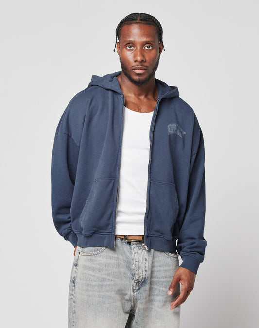 A man wears the LIVE FAST DIE YOUNG Live Fast Berlin Hooded Zip in navy blue, styled over a white tank top and light blue vintage-washed jeans, standing against a light gray background with a neutral expression.