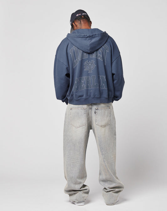 A person faces away, wearing the LIVE FAST DIE YOUNG Live Fast Berlin Hooded Zip in navy with DISTRICT BERLIN and a back graphic, light gray baggy jeans, a dark cap, all against a white background emphasizing the vintage vibe.