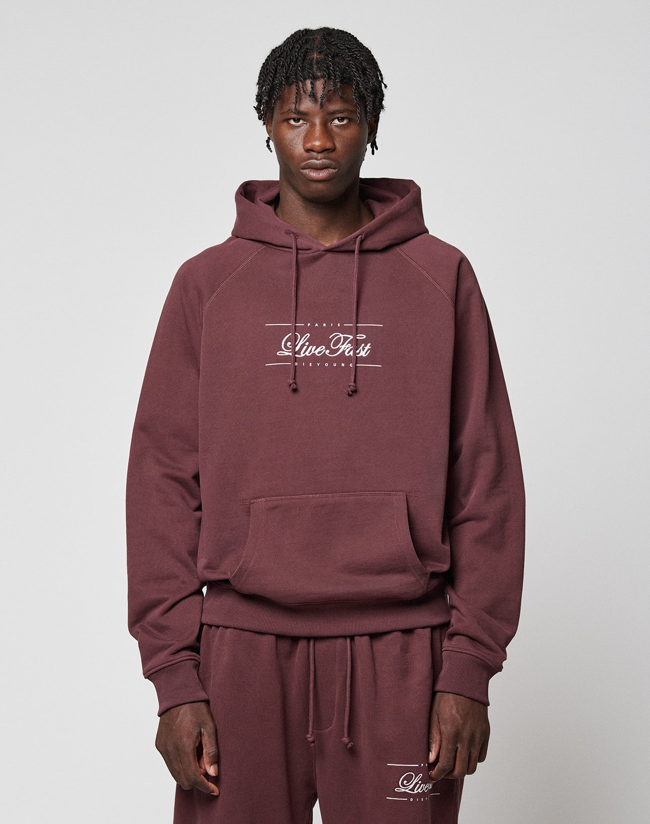 A person wears a matching maroon LFDY Embroidery Hooded and sweatpants set by LIVE FAST DIE YOUNG, crafted from soft French Terry and 100% Baumwolle. "Live Fast" is embroidered in white as they face forward against a light gray background.
