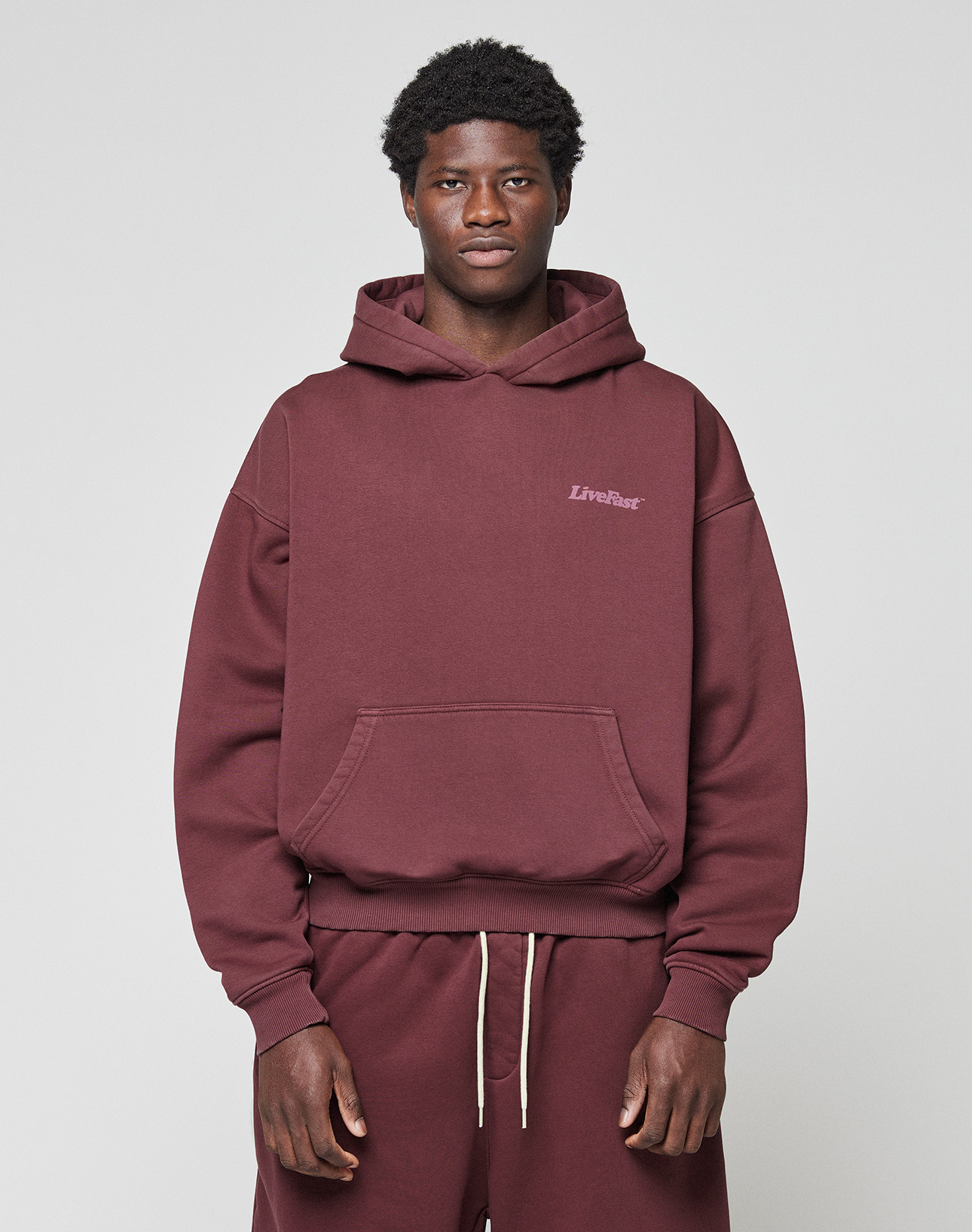 A person in an oversized maroon LIVE FAST DIE YOUNG Basic 360 Hooded sweatshirt and matching sweatpants stands facing forward against a plain light gray background. The hoodie features "LifeBag" text on the left chest.