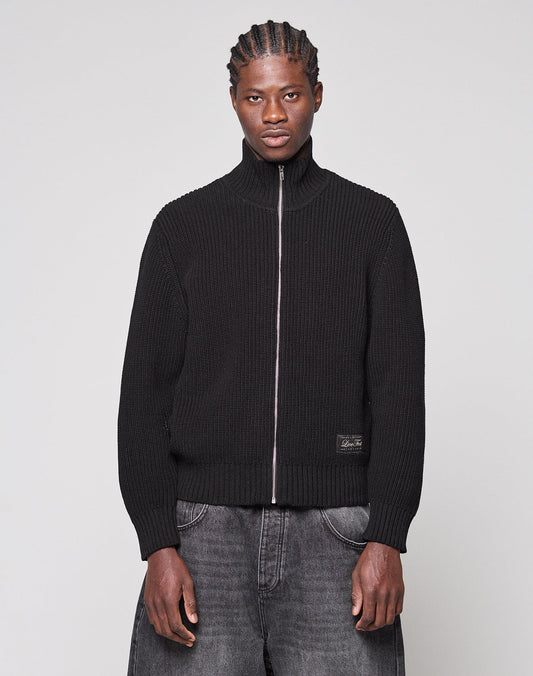 A man with braided hair models the black, ribbed LFDY Knit Jacket by LIVE FAST DIE YOUNG, featuring raglan sleeves and a stand-up collar, paired with gray washed jeans against a plain light gray background. | model