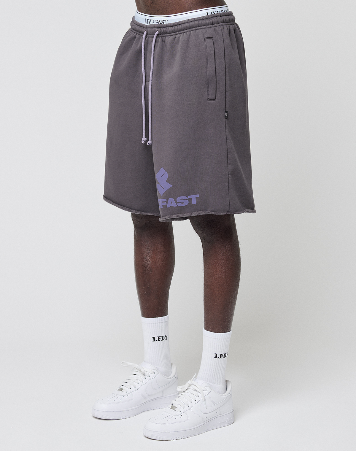A person wearing LIVE FAST DIE YOUNG Live Fast LF Shorts in gray with a purple FAST graphic, elastic waistband, side pockets, white LEDY socks, and white sneakers stands against a plain white background. | model