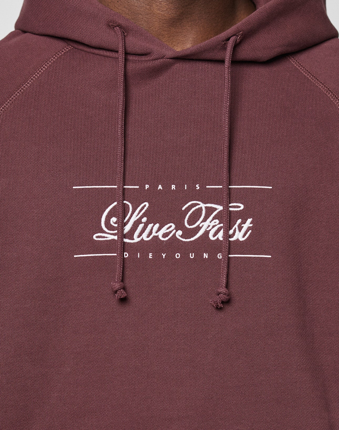 Close-up of a person wearing the LFDY Embroidery Hooded by LIVE FAST DIE YOUNG. This maroon regular fit hoodie features white embroidered text, drawstrings, and is crafted from 100% Baumwolle for comfort.