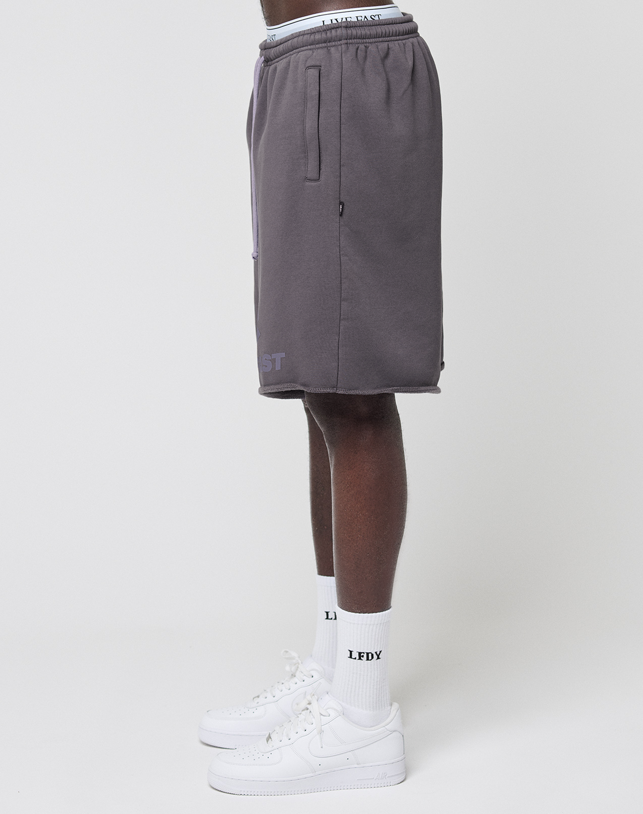 A person stands sideways wearing gray Live Fast LF Shorts by LIVE FAST DIE YOUNG, featuring an elastic waistband and side pockets, paired with white socks and sneakers. Only the lower half is visible against a plain white background.