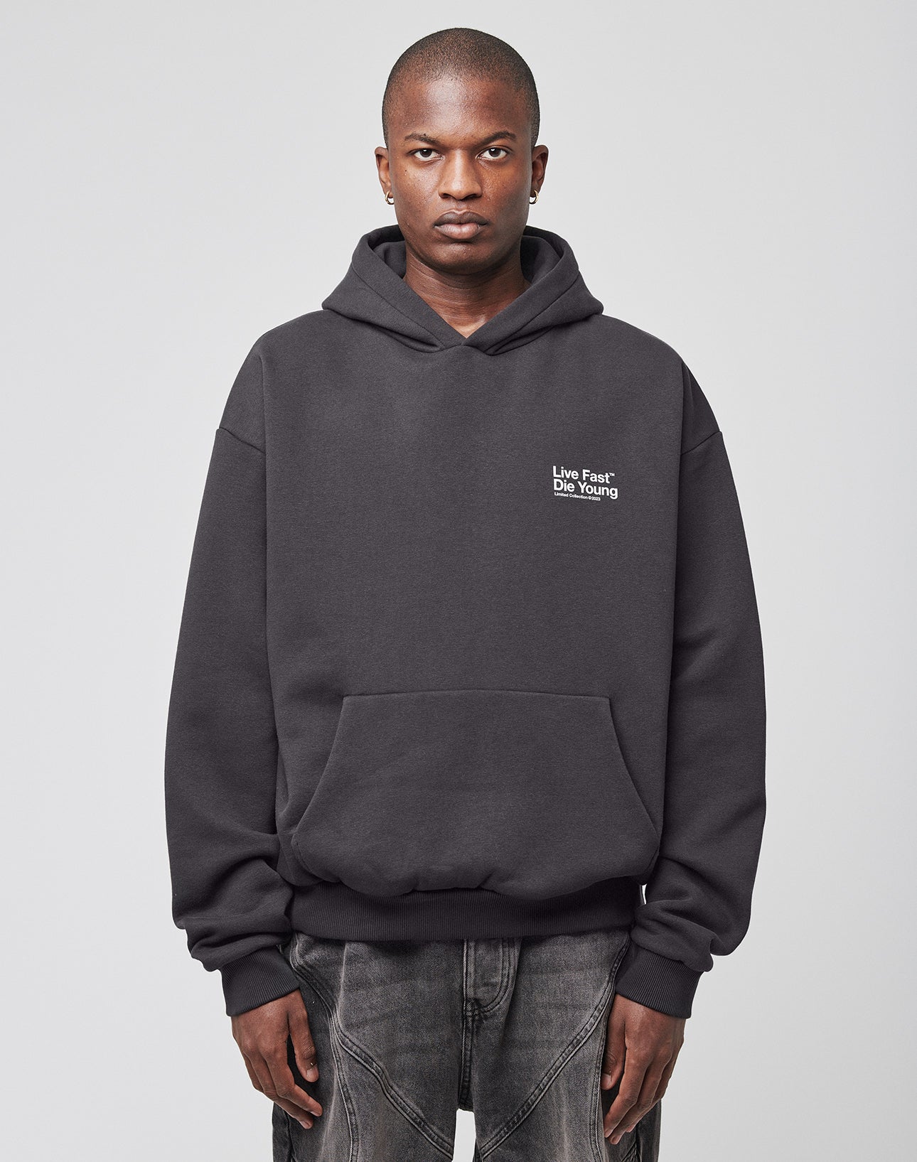 A person faces forward in an oversize fit black Live Fast NY Hooded by LIVE FAST DIE YOUNG (70% Baumwolle), featuring small white Live Fast Die Young text on the left chest, dark jeans, and a plain light background.