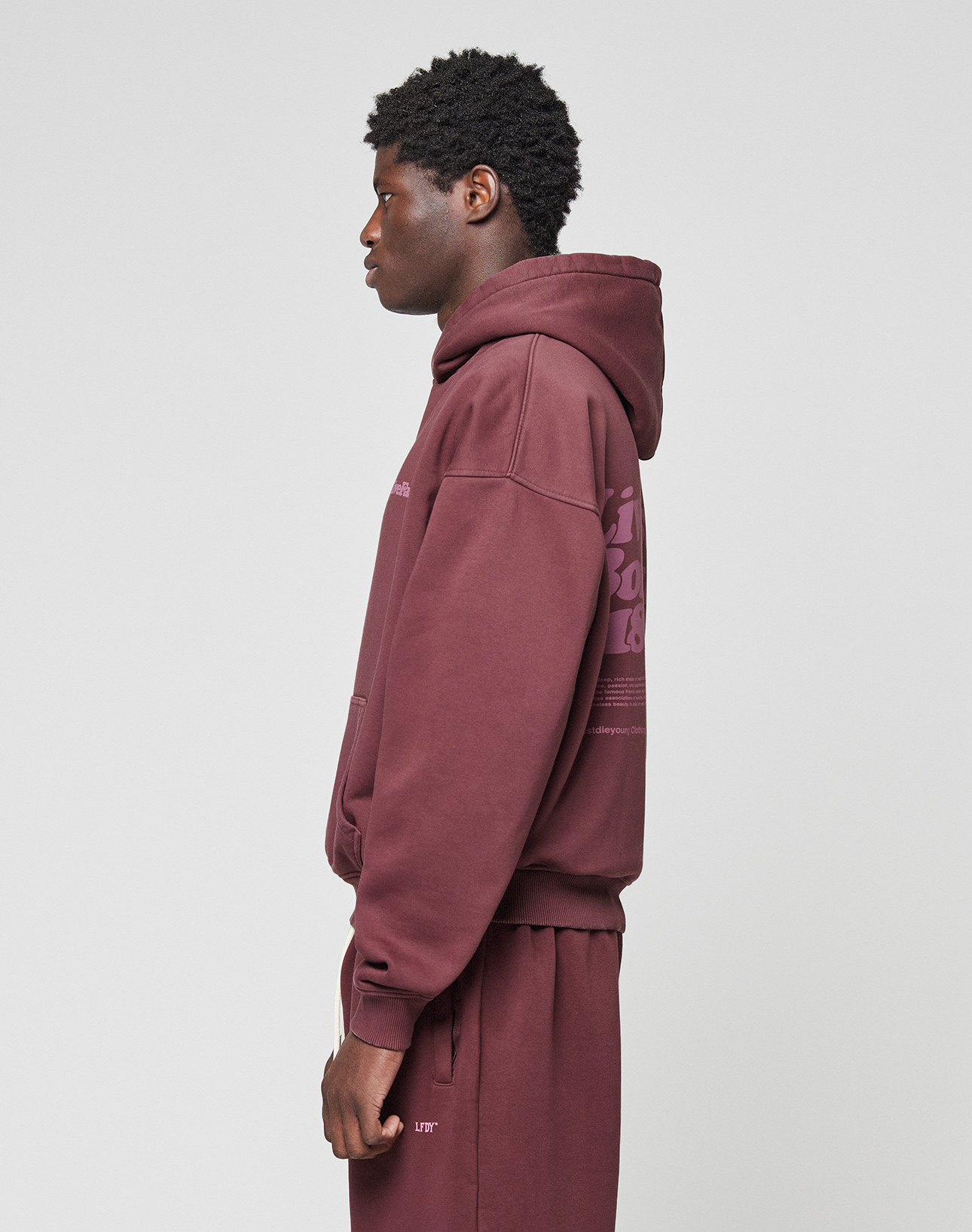 A person in an oversize fit maroon LIVE FAST DIE YOUNG Basic 360 Hooded sweatshirt and sweatpants stands in profile against a plain light gray background. The hoodie, made from weicher Baumwolle, features a hood up and subtle front and back prints.