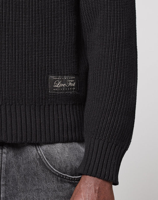 A close-up shows the lower torso and arm of a person wearing the LFDY Knit Jacket by LIVE FAST DIE YOUNG, featuring ribbed fabric, raglan sleeves, and a small "Live Fast" label near the hem, paired with faded gray jeans.