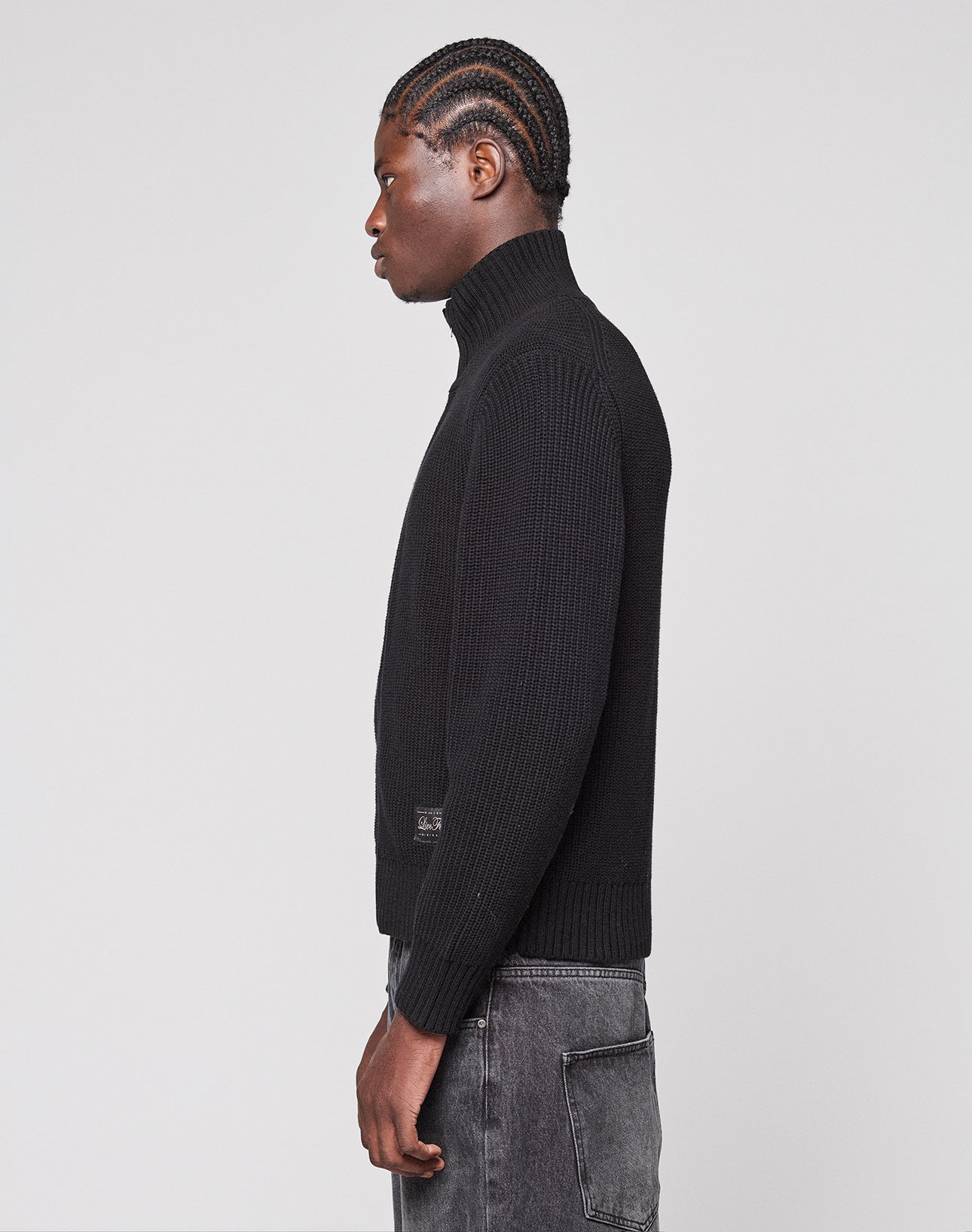 A person with braided hair stands in profile against a plain background, wearing the black LFDY Knit Jacket by LIVE FAST DIE YOUNG and dark gray jeans with a visible side pocket.