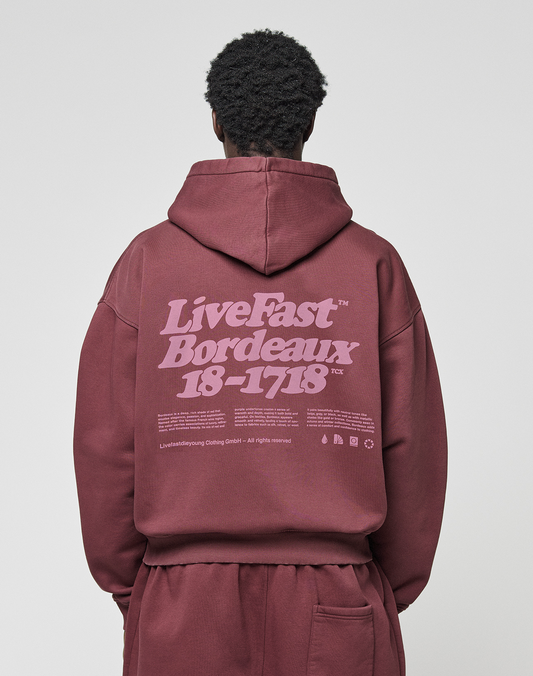 A person in an oversized maroon “Basic 360 Hooded” by LIVE FAST DIE YOUNG, featuring a bold “LiveFast Bordeaux 18-1718” backprint, stands against a plain light background facing away from the camera. | model