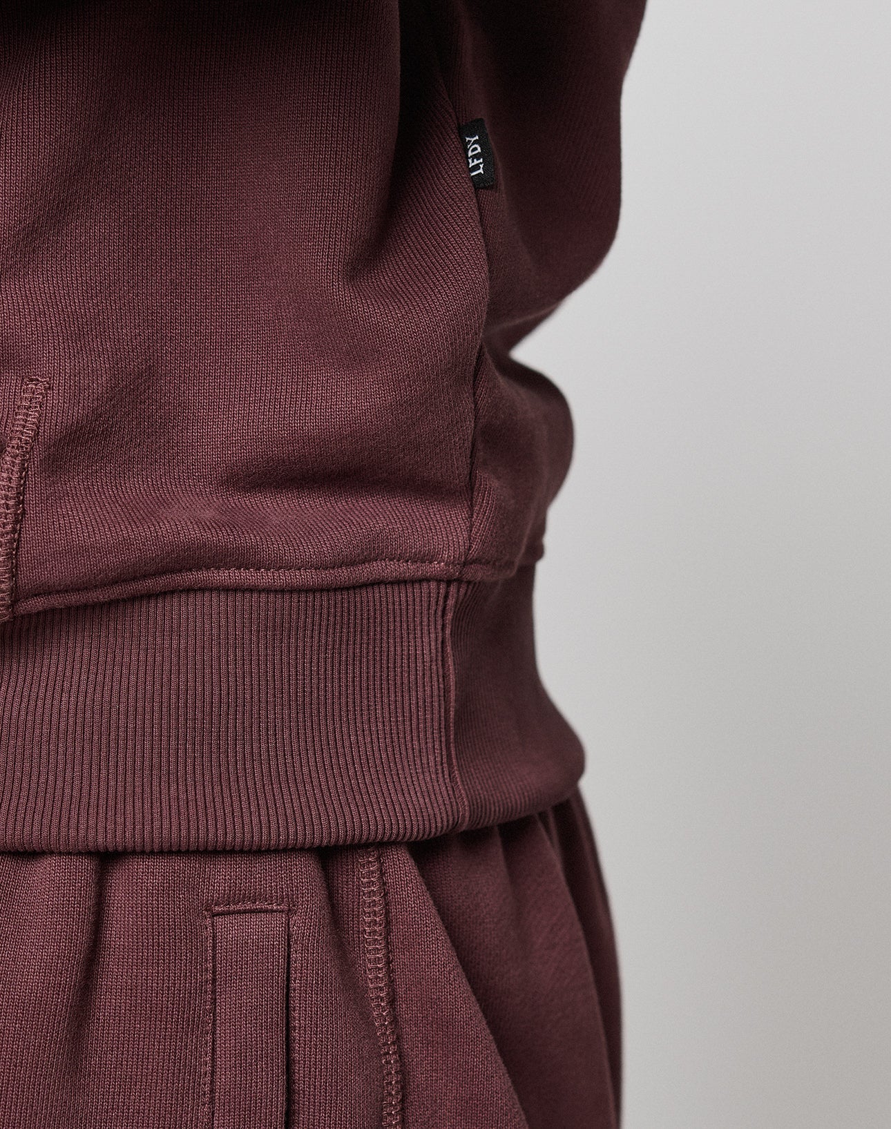 Close-up of a person in a matching maroon LIVE FAST DIE YOUNG LFDY Embroidery Hooded and sweatpants. Features include French Terry fabric, ribbed hem, drawstrings, and a small black tag on the 100% Baumwolle sweatshirt.