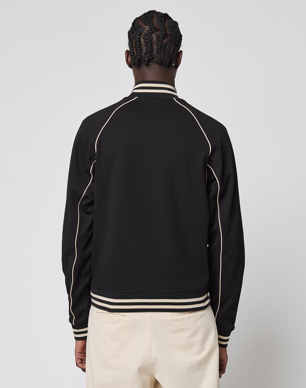 A person with braided hair, seen from the back, wears the Live Fast Tracksuit Jacket—black with white piping, Chainstitch Logo, striped collar and cuffs, Eingrifftaschen—paired with light pants against a plain light background.