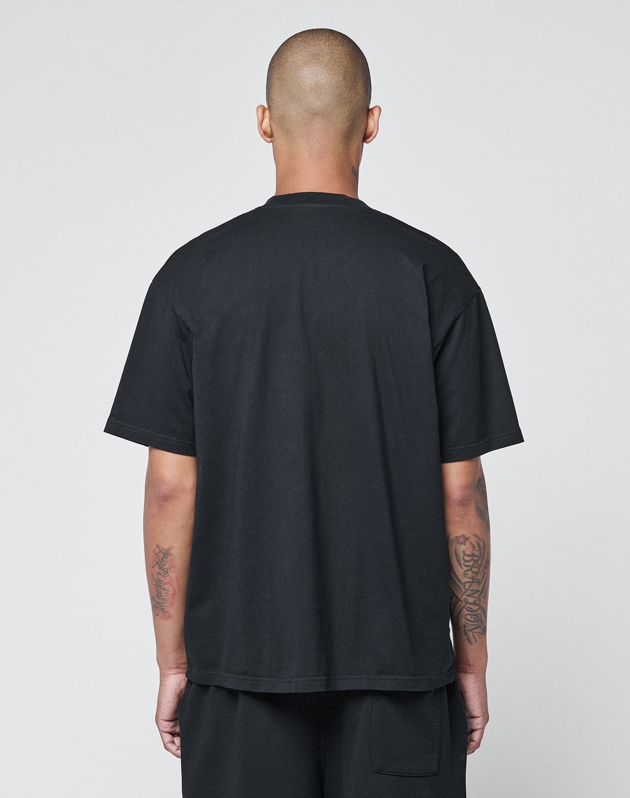 A person with a shaved head faces away from the camera, wearing the LIVE FAST DIE YOUNG Basic Chest Tee in black and shorts. Their tattooed forearms are visible, arms by their sides, against a plain light background.