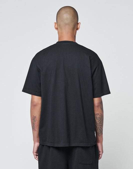 A person with a shaved head faces away from the camera, wearing the LIVE FAST DIE YOUNG Basic Chest Tee in black and shorts. Their tattooed forearms are visible, arms by their sides, against a plain light background.