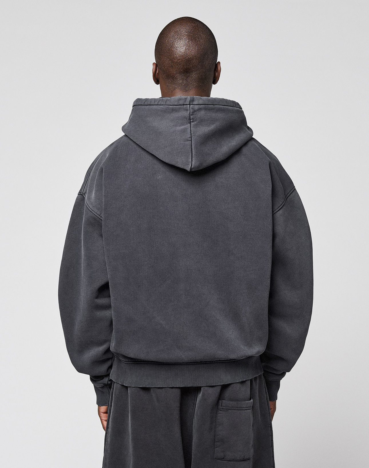 A person with a shaved head stands with their back to the camera, wearing the LFDY 40 Hooded oversize-fit dark gray sweatshirt and matching pants by LIVE FAST DIE YOUNG. The set features a kangaroo pocket and is made from cotton-polyester. The background is plain and light.