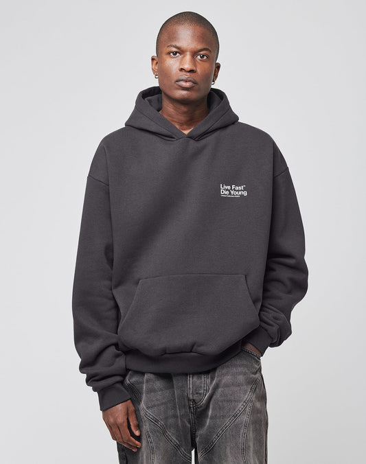 A person stands facing the camera in an oversize fit Live Fast NY Hooded by LIVE FAST DIE YOUNG, featuring a Live Fast Die Young front print and faded jeans, against a plain light background. The hoodie is made from 70% Baumwolle for comfort.
