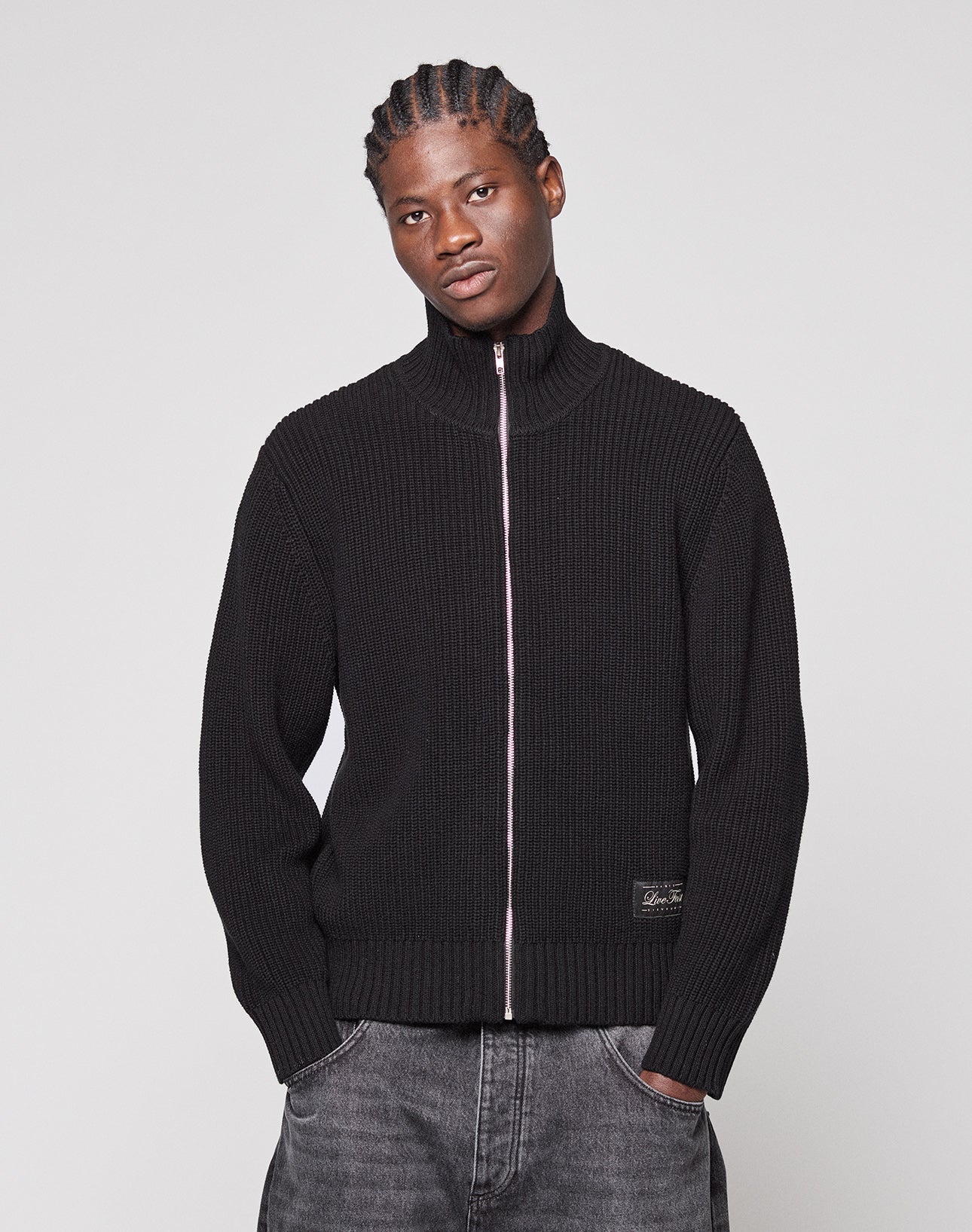 A person with braided hair wears the black LFDY Knit Jacket by LIVE FAST DIE YOUNG—100% cotton, ribbed with zip-up front and pockets—paired with dark jeans, standing against a plain light gray background.