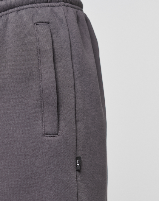 Close-up of a person wearing Live Fast LF Shorts by LIVE FAST DIE YOUNG in dark gray with side pockets, wide fit, and a small black LOUT tag near the pocket. An elastic waistband offers comfort. Background is plain and light-colored.