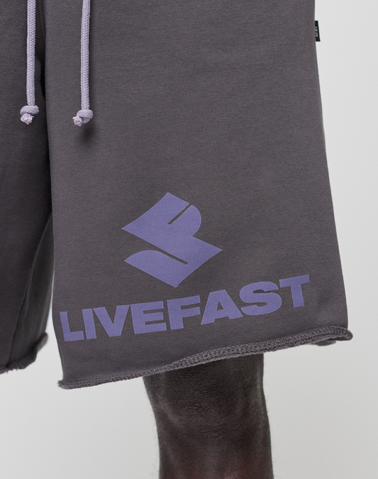 Close-up of a person wearing LIVE FAST DIE YOUNG Live Fast LF Shorts in dark gray, featuring a purple geometric logo and LIVEFAST text on the left leg, wide fit with visible elastic waistband, raw edges, and practical side pockets.