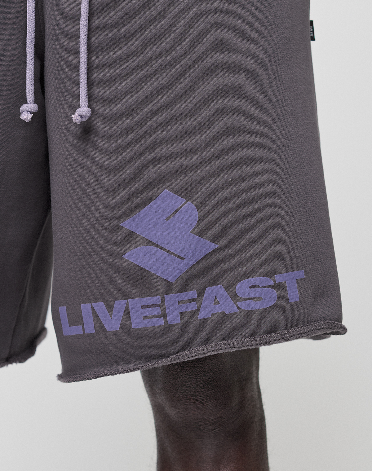 Close-up of a person wearing LIVE FAST DIE YOUNG Live Fast LF Shorts in dark gray, featuring a purple geometric logo and LIVEFAST text on the left leg, wide fit with visible elastic waistband, raw edges, and practical side pockets.
