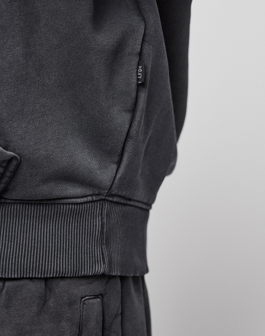 Close-up of a person wearing a dark gray LFDY 40 Hooded sweatshirt by LIVE FAST DIE YOUNG with matching pants. A small fabric tag is visible on the side seam, and the top features a subtle kangaroo pocket. The background is plain and light-colored.