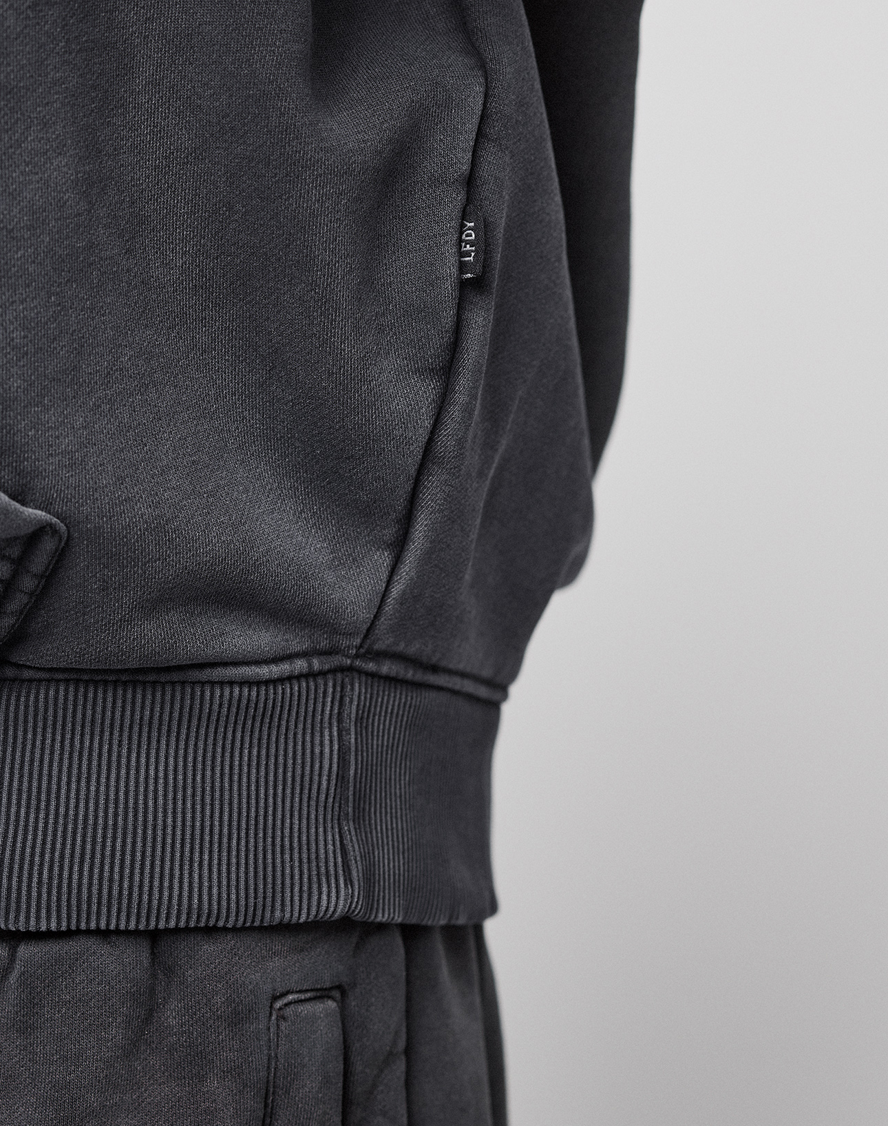 Close-up of a person wearing a dark gray LFDY 40 Hooded sweatshirt by LIVE FAST DIE YOUNG with matching pants. A small fabric tag is visible on the side seam, and the top features a subtle kangaroo pocket. The background is plain and light-colored.
