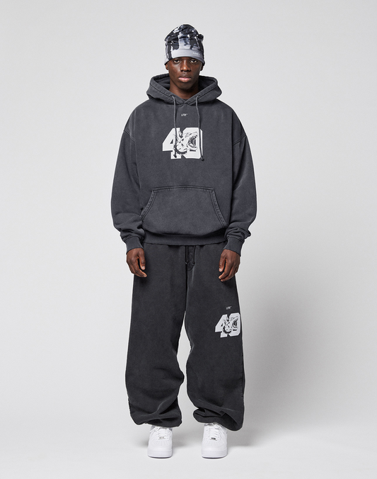 A man faces forward wearing the LIVE FAST DIE YOUNG LFDY 40 Hooded in dark gray with white graphics and a Kangaroo-Tasche, paired with matching baggy sweatpants, a patterned headscarf, and white sneakers against a plain light gray background.
