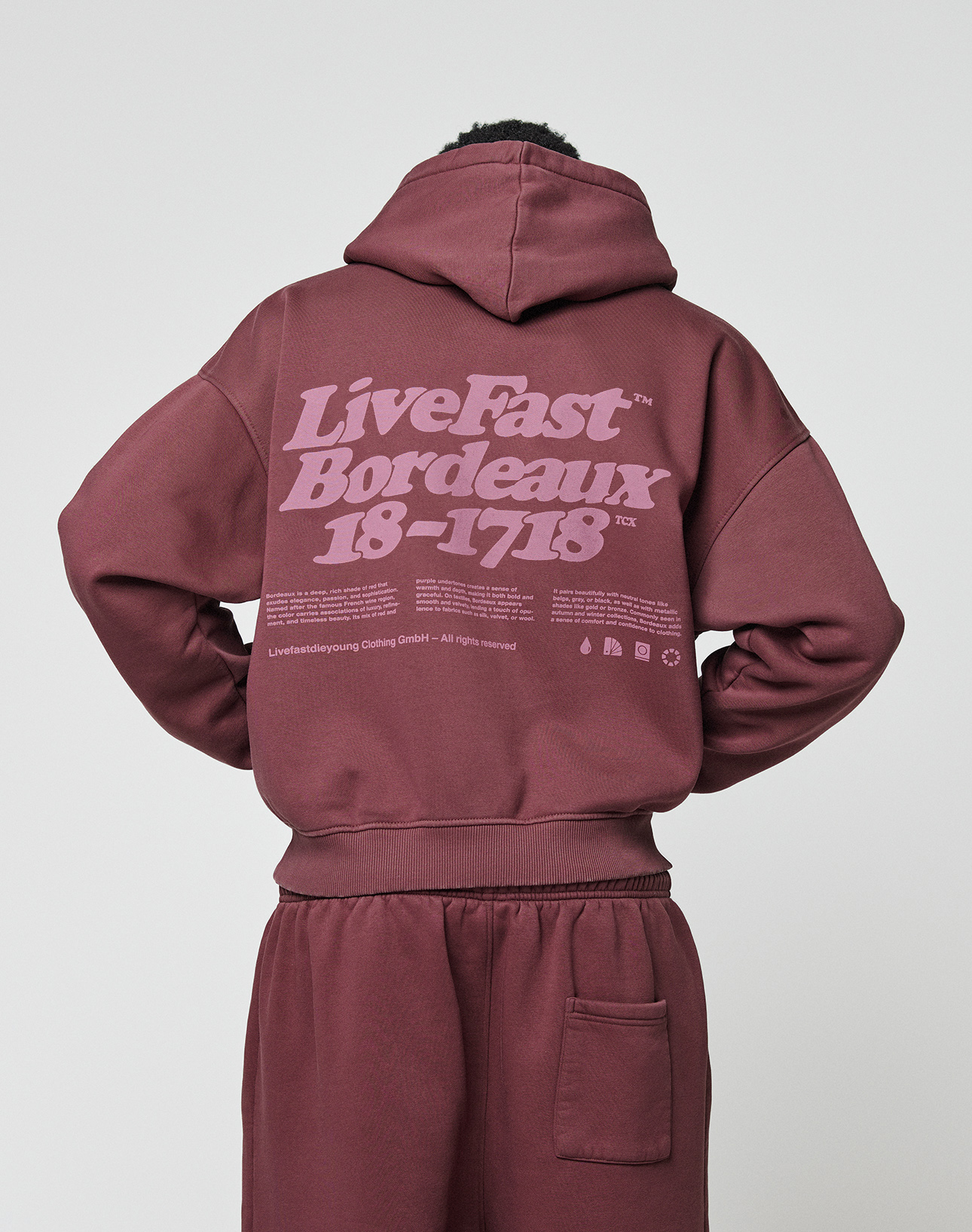A person in a maroon Basic 360 Hooded and matching pants by LIVE FAST DIE YOUNG, seen from the back. The hoodie features large pink "LiveFast Bordeaux 18-1718" text and additional smaller print on the front and back.