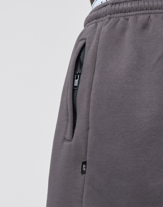 Close-up of a person wearing LIVE FAST DIE YOUNG's Live Fast LF Shorts in dark gray, featuring elastic waistband, wide fit, side slit pockets with a zippered pocket, and a small black tag with white text near the seam.