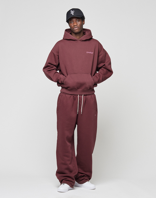 A person stands against a light gray background wearing the LIVE FAST DIE YOUNG Basic 360 Hooded maroon oversize hoodie and sweatpants set, with hands in the hoodie pocket, plus a black baseball cap and white sneakers.