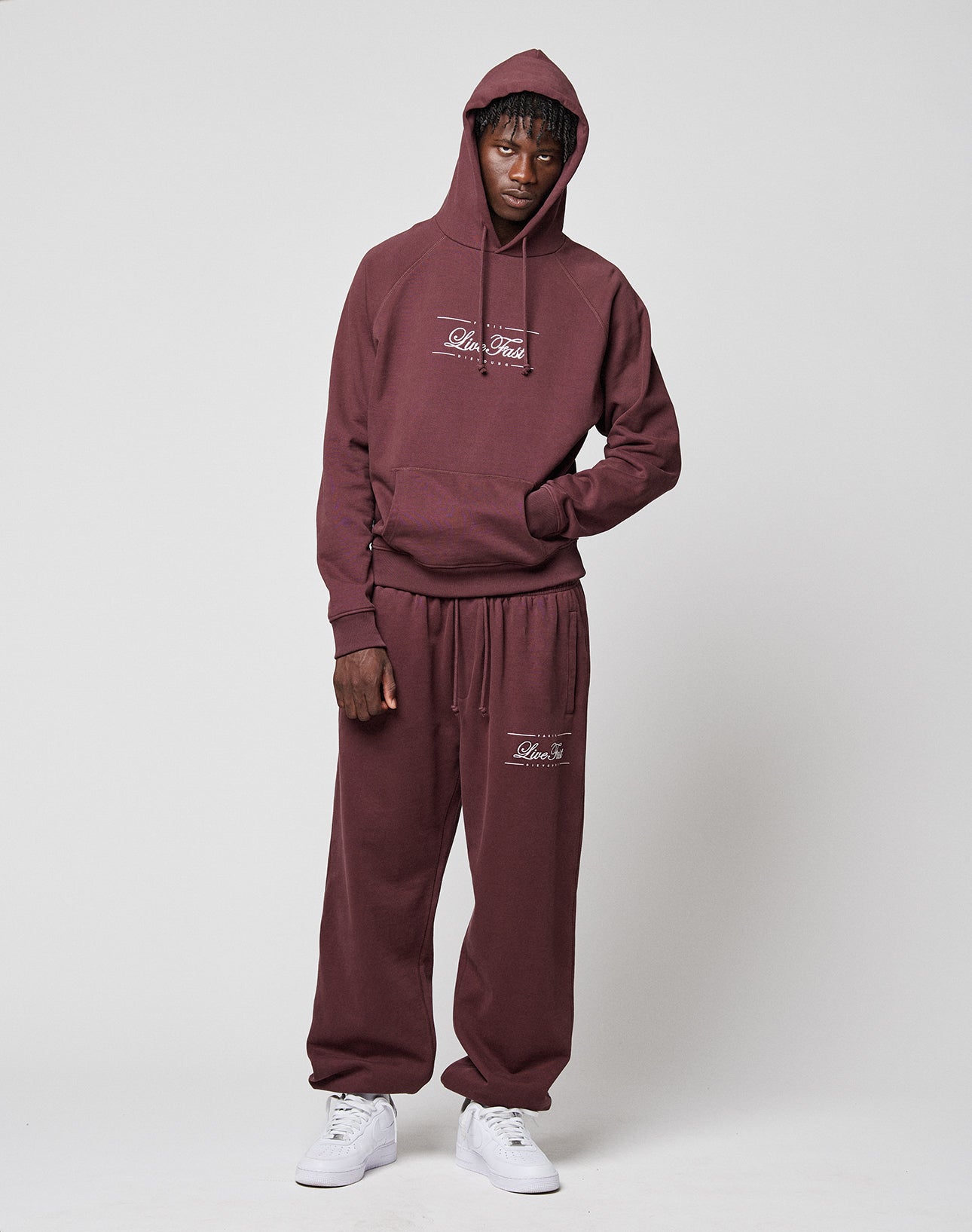 A person stands against a plain background wearing the LIVE FAST DIE YOUNG LFDY Embroidery Hooded and matching sweatpants set in maroon, both featuring white script logos. The 100% Baumwolle hoodie is worn with the hood up, one hand in the pocket, paired with white sneakers.