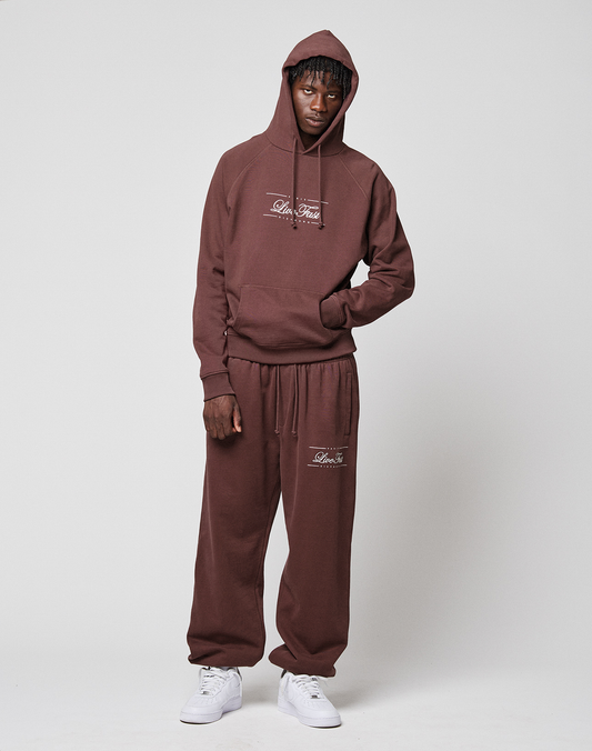 A person stands against a plain background wearing a matching brown French Terry hoodie and LIVE FAST DIE YOUNG Wide Cuff Jogger with white lettering and sneakers. One hand is in their pocket as they look at the camera.