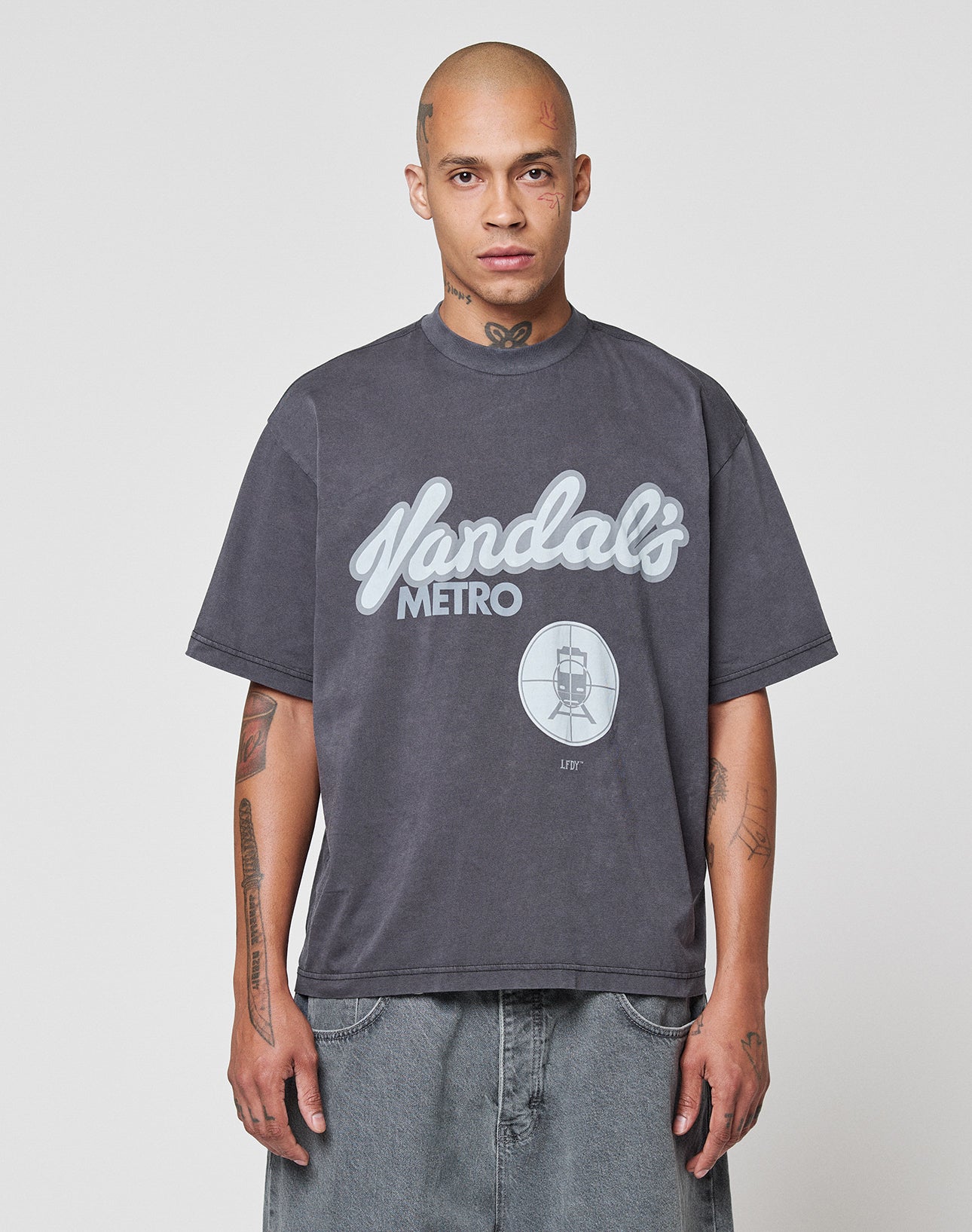 A person with a shaved head and tattoos wears the LIVE FAST DIE YOUNG LFDY Vandals Tee—an oversize fit, 100% cotton dark gray shirt with a Metro frontprint and train graphic—paired with baggy denim shorts against a white background. | model