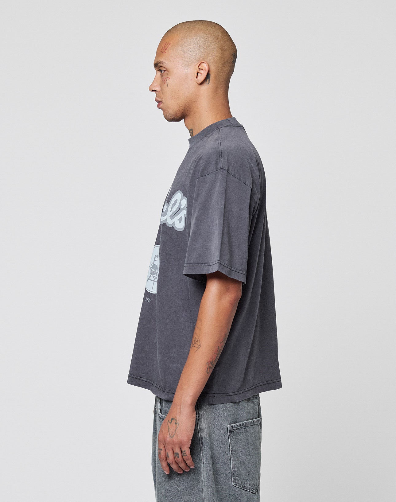 A person with a shaved head stands in profile, wearing the LIVE FAST DIE YOUNG LFDY Vandals Tee, a loose-fitting, faded gray graphic T-shirt made of 100% Baumwolle, paired with light gray jeans against a plain light background.