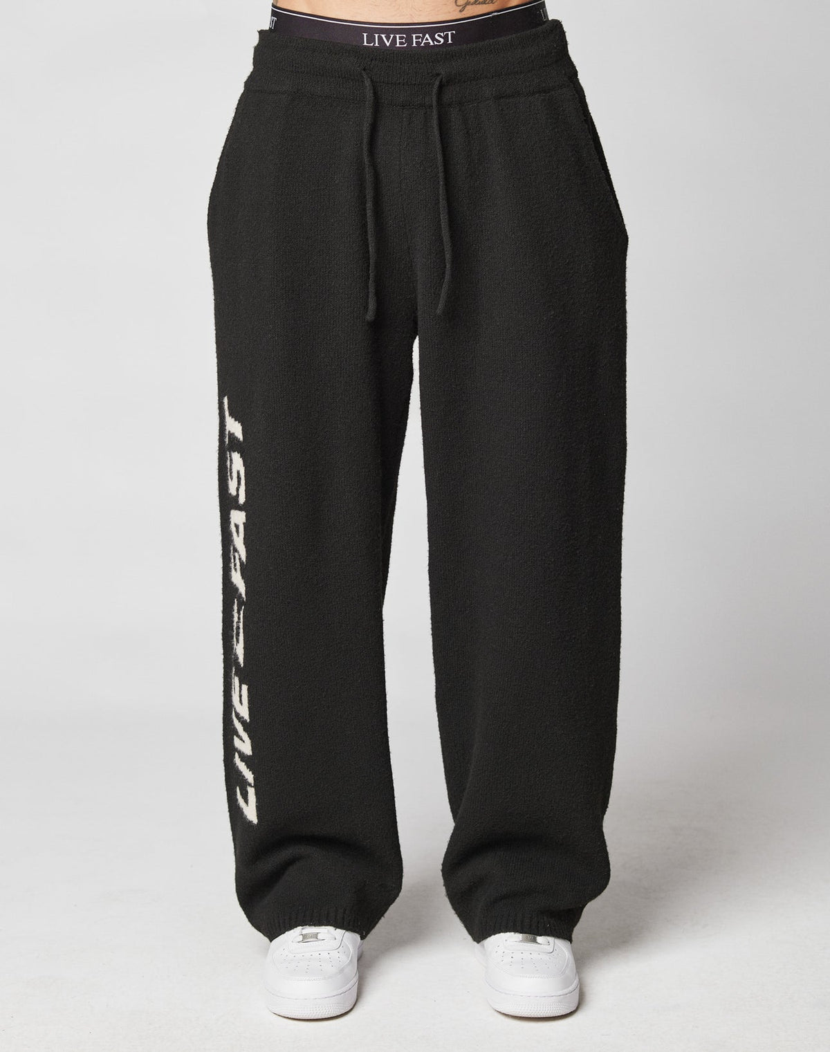 A person wears LIVE FAST DIE YOUNG’s Live Fast Boucle Pants—wide fit, black, with LIVE FAST in white down one leg and on the elastic waistband—paired with white sneakers. 100% polyester for comfort and style.