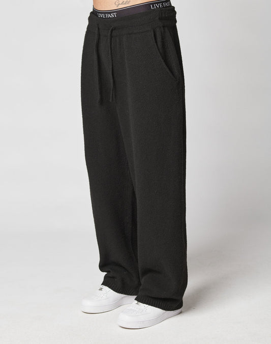 A person wears LIVE FAST DIE YOUNG Live Fast Boucle Pants—black, wide-fit, 100% polyester sweatpants with an elastic waistband printed LIVE FAST and side pockets, styled with white sneakers against a plain light background.
