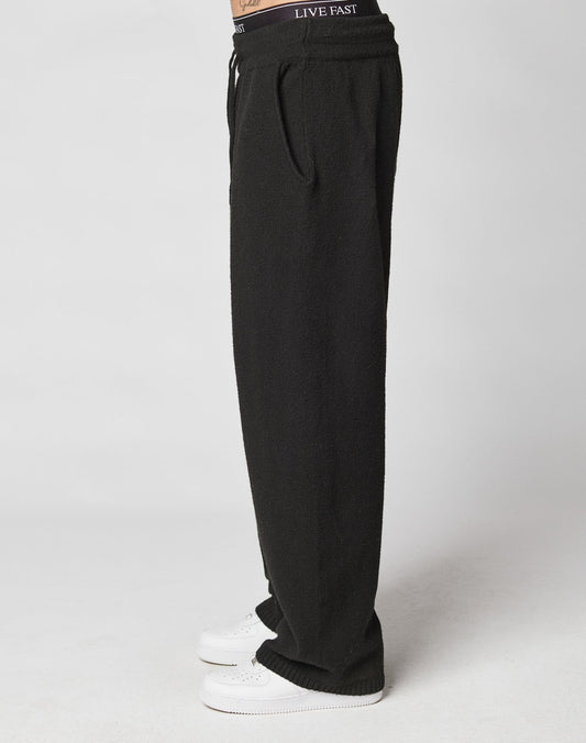 A person wearing wide fit, loose-fitting black Live Fast Boucle Pants by LIVE FAST DIE YOUNG with an Elastik-Bund and white sneakers stands sideways. The pants waistband reads LIVE FAST; the background is plain and light-colored.